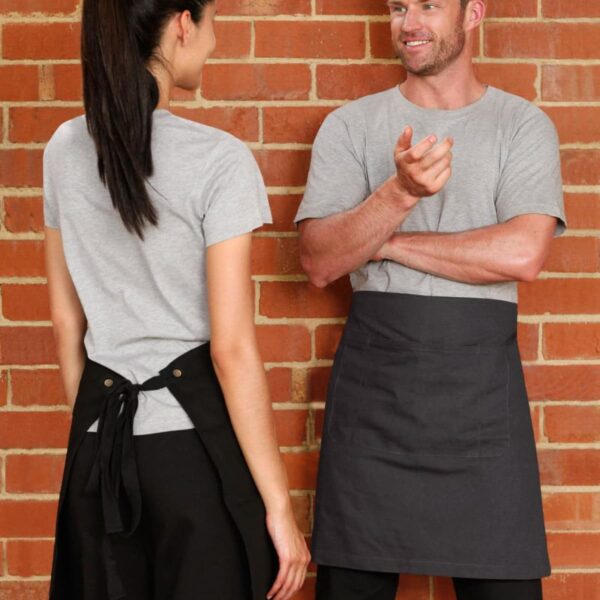 Two people wearing gray shirts and black aprons are having a conversation in front of a brick wall. One person is facing the other with arms folded, while the other stands with their back partially turned.