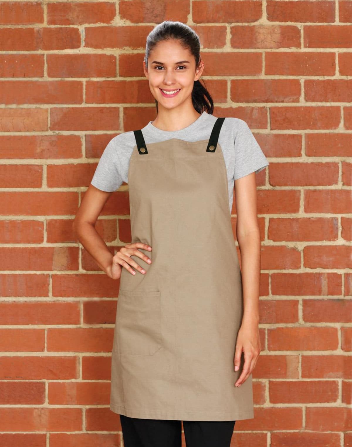 Person in beige apron and gray shirt smiles in front of a brick wall.
