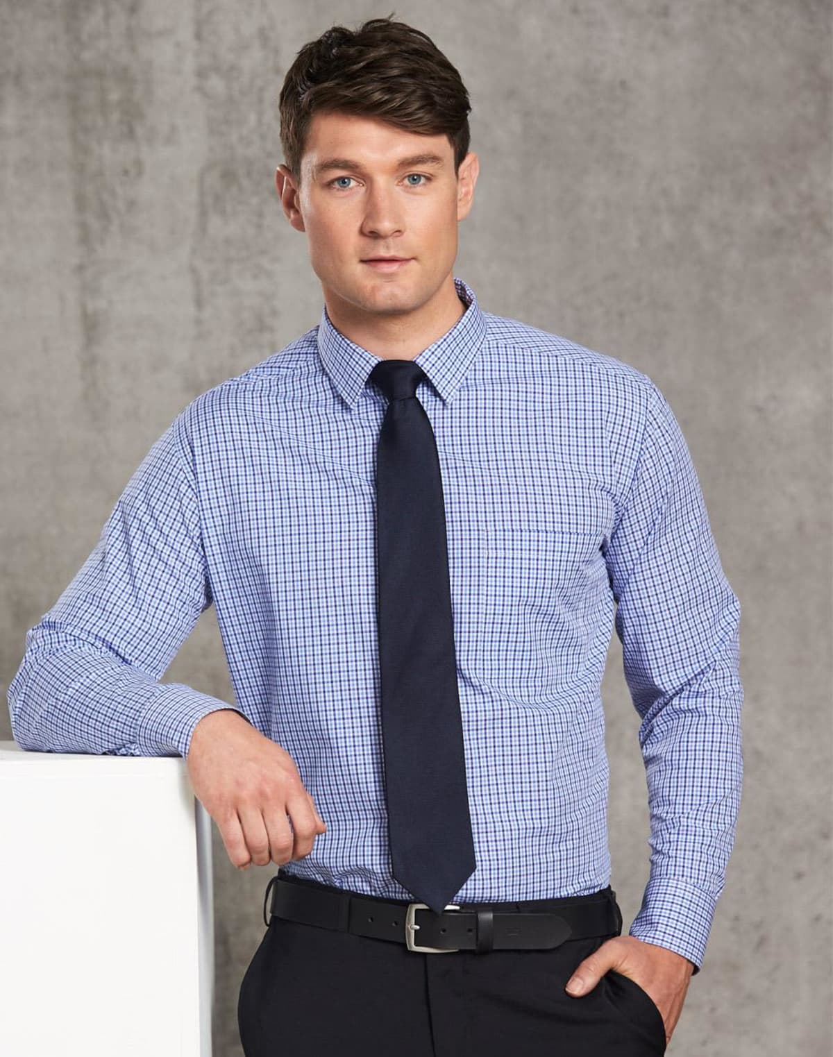 Man in a blue checkered shirt and dark tie, standing against a gray background.