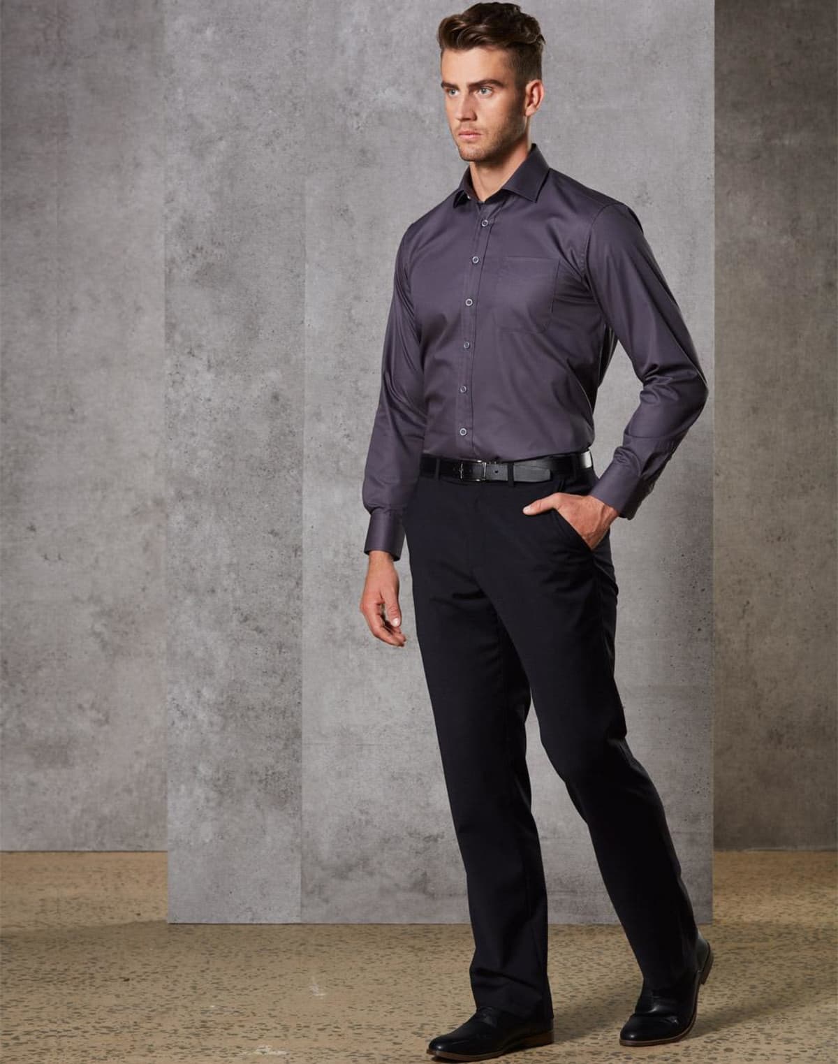 Man in a dark shirt and trousers standing against a gray wall.