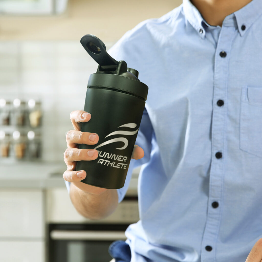 Person holding a dark green Runner Athlete protein shaker in a kitchen.