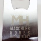 Metal bottle opener card with a cut-out opener at the top. It features the text MH Maschler Homes and a phone number engraved below. The design includes a small hole in the bottom left corner.
