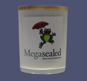 White candle with wooden lid, featuring a green frog holding a red umbrella and the text Megasealed Bathrooms & Balconies.