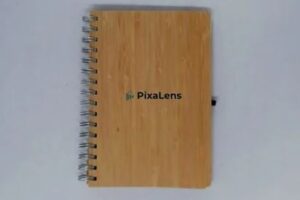 Wooden spiral notebook with PixaLens on the cover, placed on a white background.