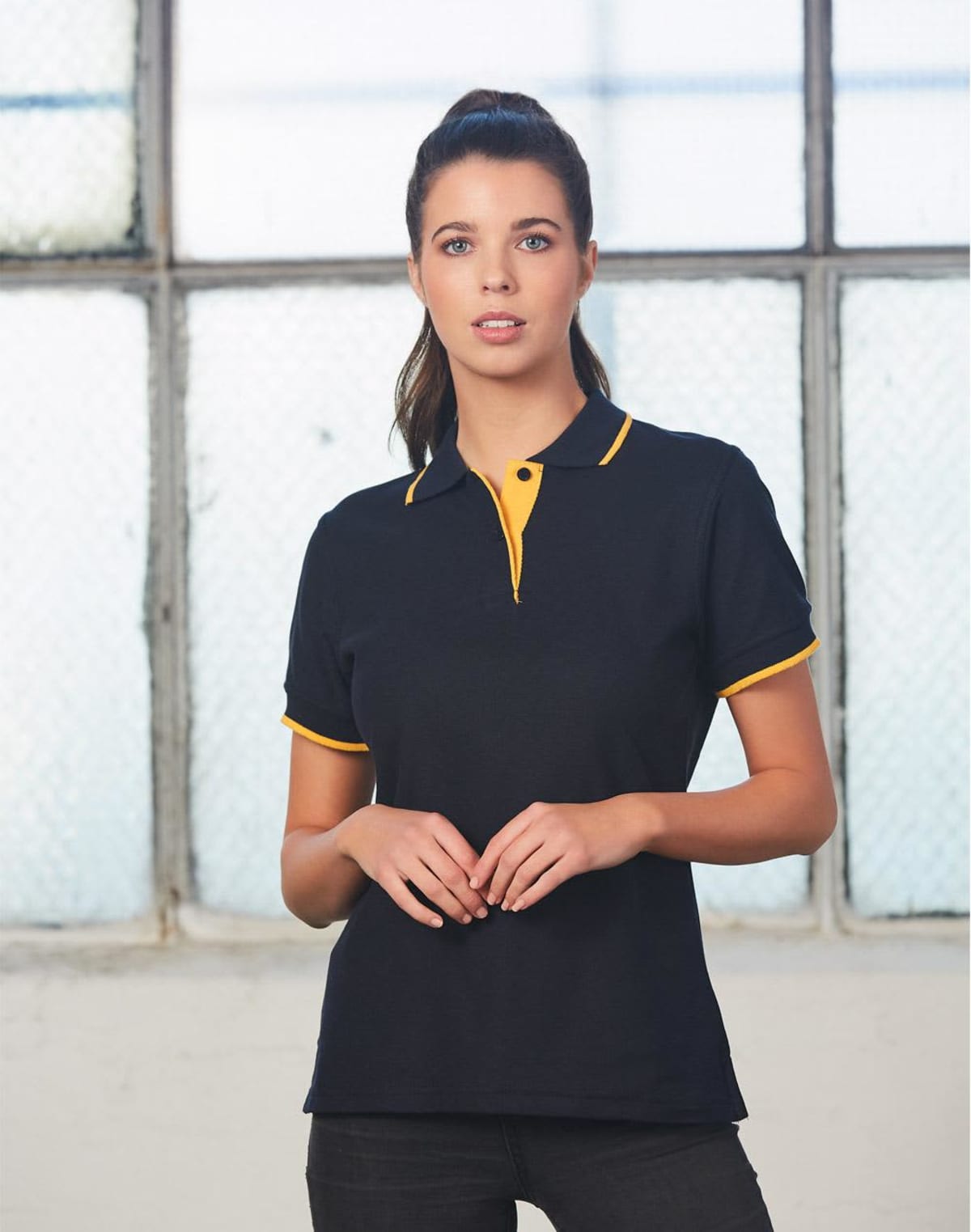 Person wearing a black polo shirt with yellow trim, standing in front of a textured window.