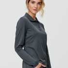 Woman in a dark gray long-sleeve polo shirt smiles slightly, hand in pocket.