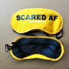 Two yellow sleep masks on a brown surface. The top mask displays the text SCARED AF #GCFILMFEST in black and white letters. The bottom mask is plain with black trim and straps.