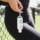 Hand holding a bottle of hand sanitizer clipped to pants, near a chain-link fence outdoors.