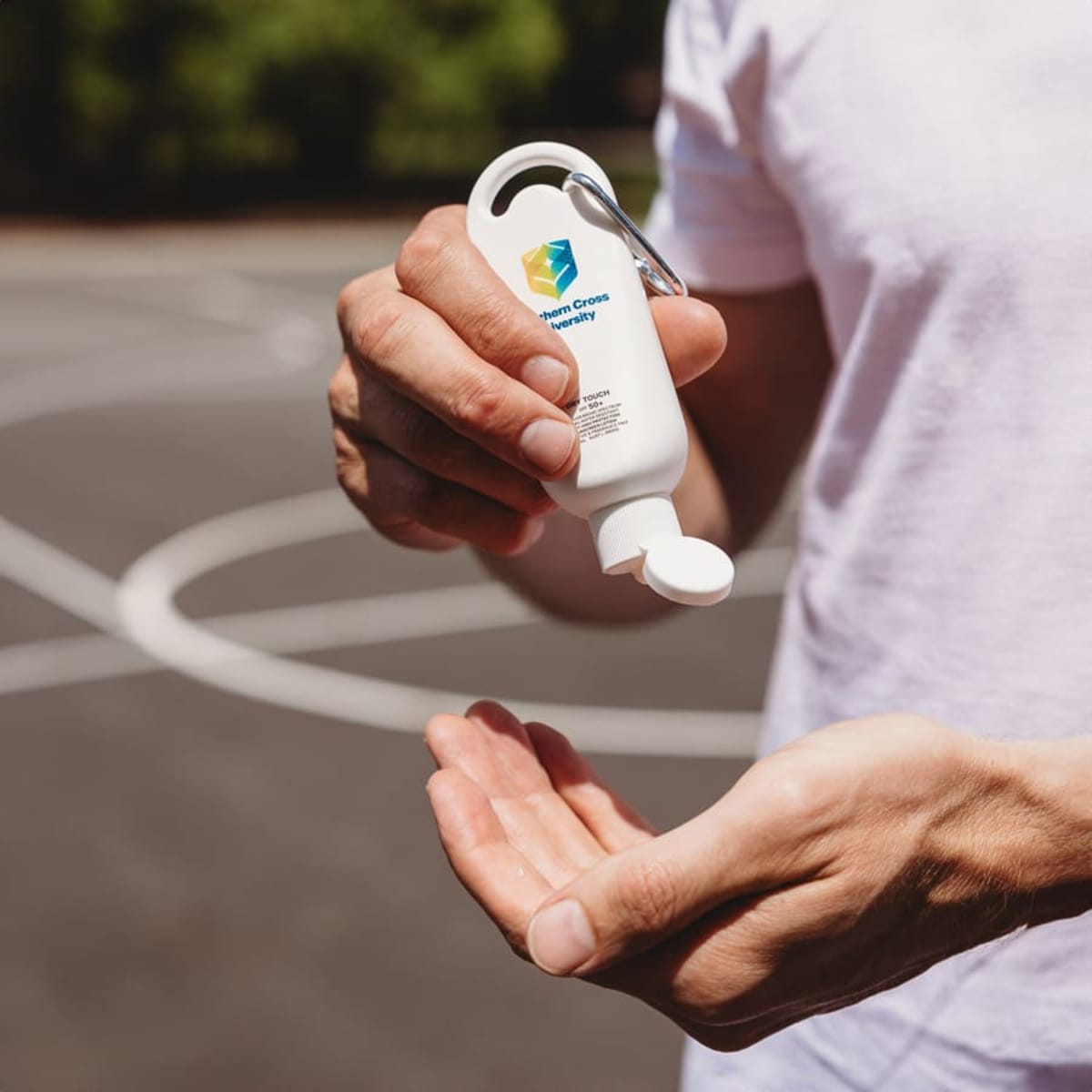 Person applying sunscreen from a small bottle on a basketball court outdoors.
