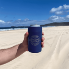A hand holding a blue beverage holder with a can inside, labeled Skinny Creek Brewery with a logo. The scene is a sunny beach with clear skies, sand, and distant hills.