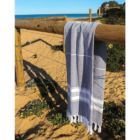 A striped beach towel hangs on a wooden fence by a sandy beach with greenery nearby.