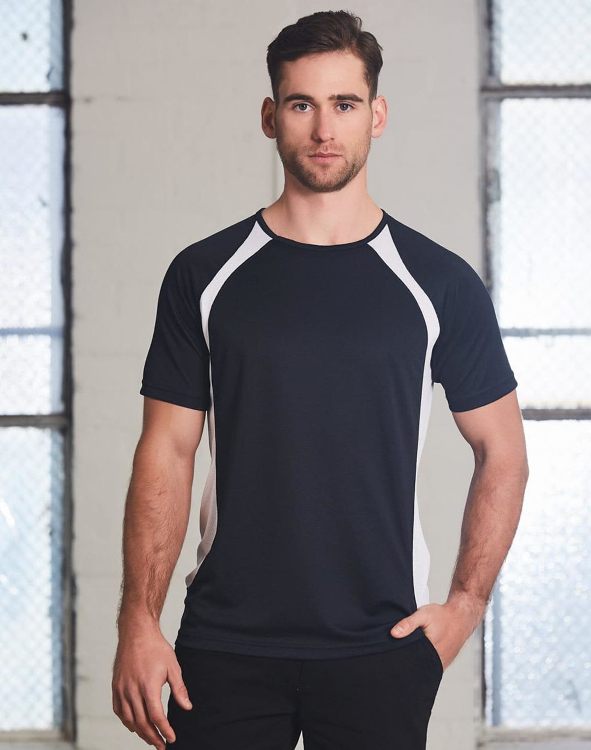 Man in black and white t-shirt stands against a textured wall with windows.