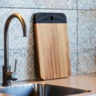 Cutting board leaning against a patterned tile wall above a metal kitchen sink and faucet.