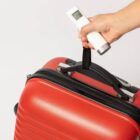 A hand holds a luggage scale measuring the weight of a red suitcase.