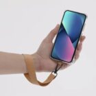 Hand holding a smartphone with a brown wrist strap attached to the phone case.