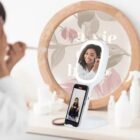 Woman applies makeup in front of a smart mirror with a phone attached, reflecting her smile.