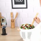 Person stirring salad while filming with a smartphone on a stand in a kitchen.