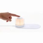 A hand touches a small, glowing lamp on a white charging pad.