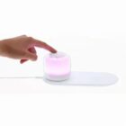 A hand touches a small glowing pink device on a white base with a wire attached.