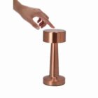 A hand reaches to touch a modern, copper-colored table lamp on a white background.