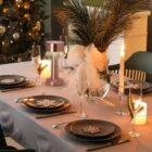 Elegant holiday table set with black plates, candles, and festive decorations near a lit tree.