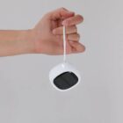 A hand holds a small white device with a black front panel and a white string loop.