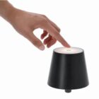 Hand reaching to touch the top of a small, black, conical lamp with a lighted surface.