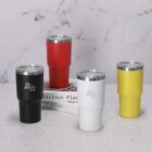 Four colored travel mugs on a counter with books in the background.