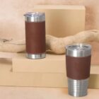 Two brown travel mugs with silver tops on beige blocks, wood branch in background.