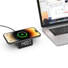 Smartphone charging on a wireless dock with a digital clock, next to a laptop on a desk.