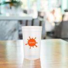 Clear cup with an orange Sports splash logo on a wooden table.
