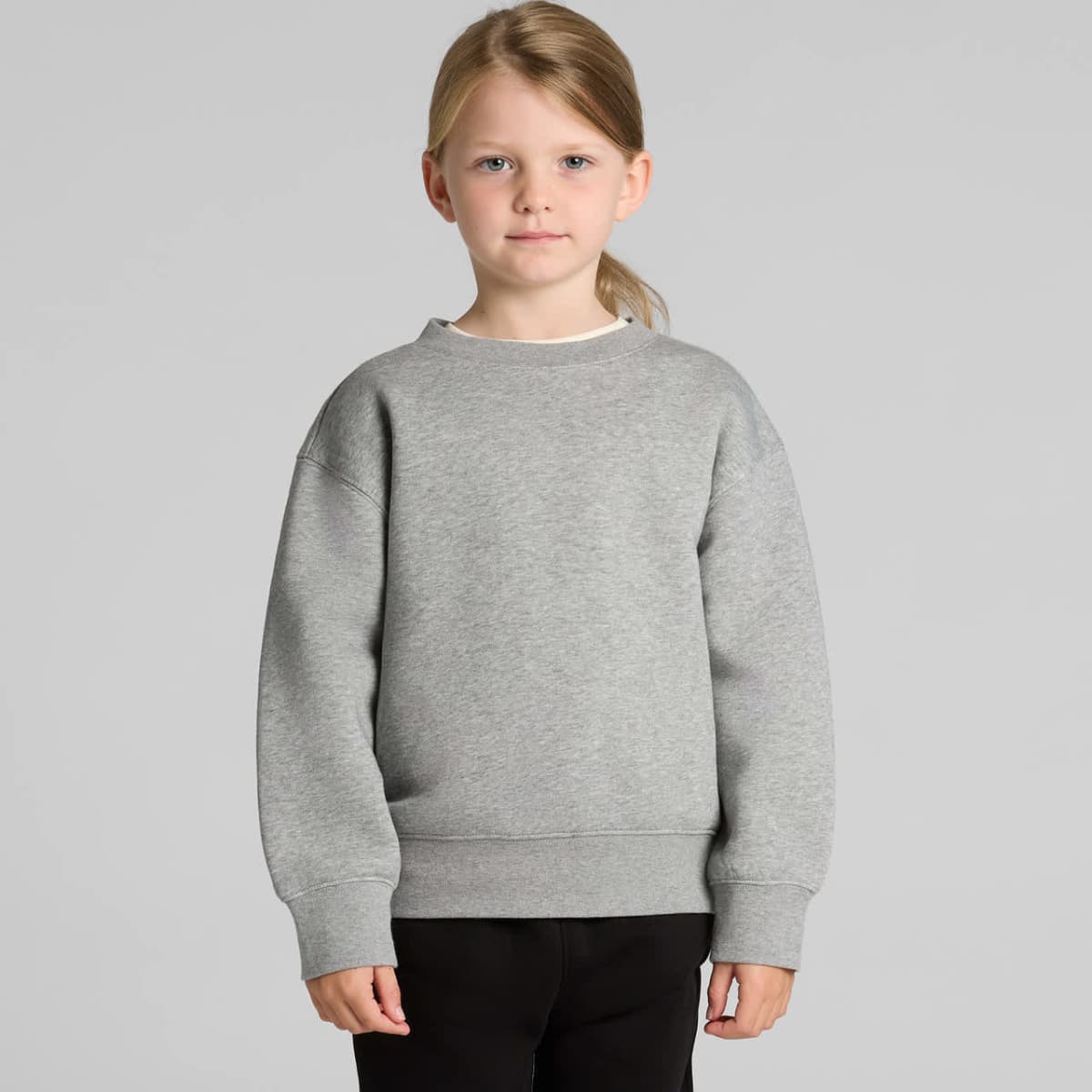 Young girl in a gray sweater and black pants standing against a plain background.