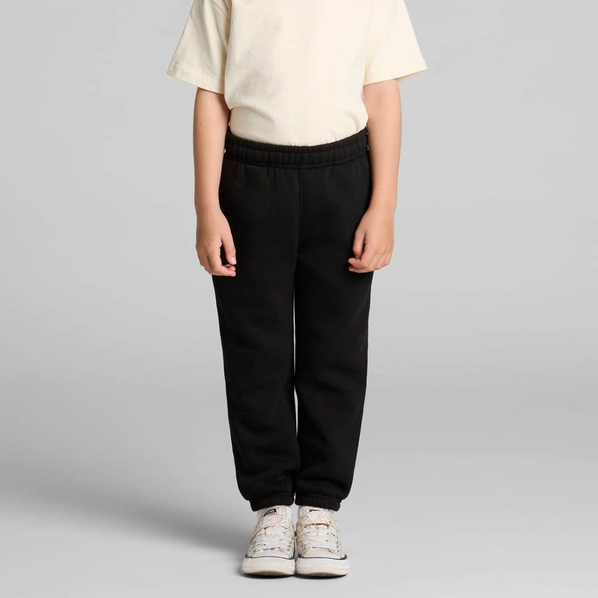 Child wearing a cream shirt, black pants, and white sneakers against a gray background.