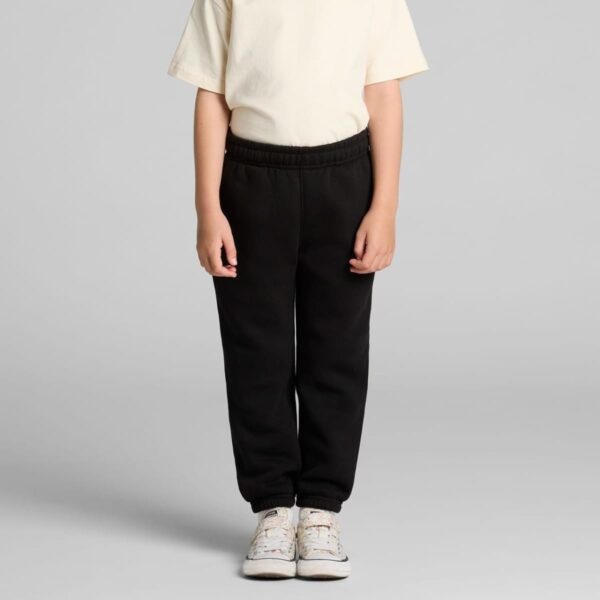 Child wearing a cream shirt and black pants, standing against a gray background.