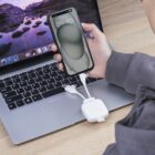 Person using smartphone connected to a laptop and wireless earphones on a wooden table.