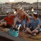 Friends toasting with drinks on a rooftop, wearing party hats, with a wine bucket nearby.