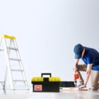 Person using drill on wooden plank; ladder and toolbox nearby.