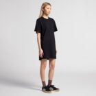 Person in black T-shirt dress, brown socks, and black sneakers stands against a plain background.