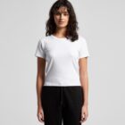 Woman with wavy hair wearing a plain white t-shirt and black pants, standing against a gray background.