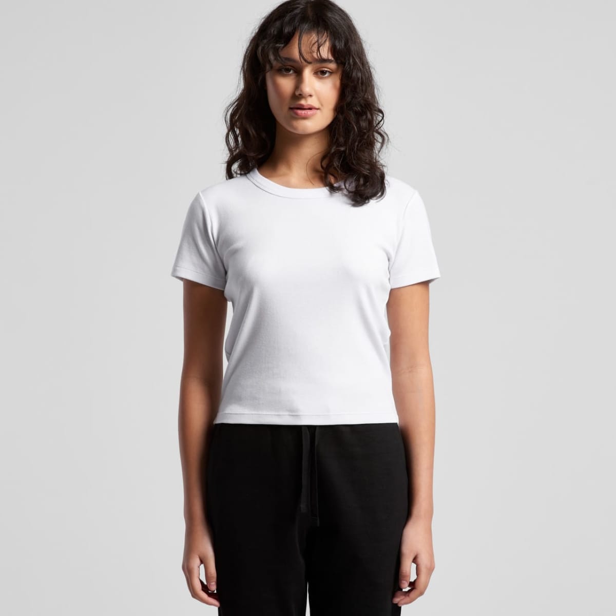 Woman with wavy hair wearing a plain white t-shirt and black pants, standing against a gray background.