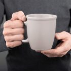 A person holding a plain white mug with both hands, wearing a dark long-sleeve shirt.