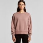 Woman in a mauve sweatshirt and black pants standing against a plain gray background.