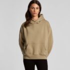 Woman in a tan hoodie and black pants standing against a plain light gray background.