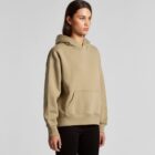 A woman wearing a beige hoodie and black pants stands against a plain light background.