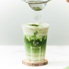 A matcha latte in a clear cup with powder being sifted on top.