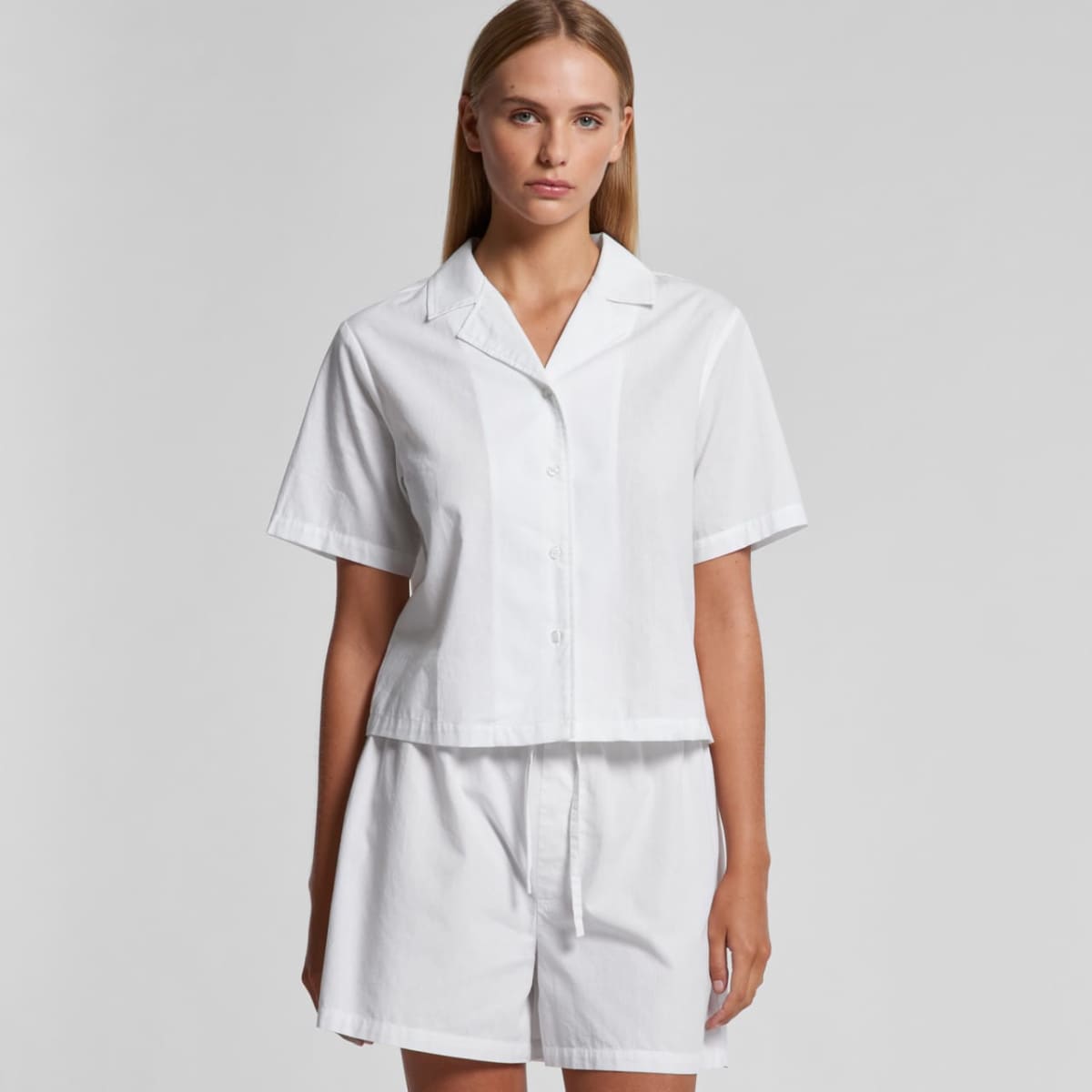 Woman in white short-sleeve button-up shirt and shorts against a gray background.