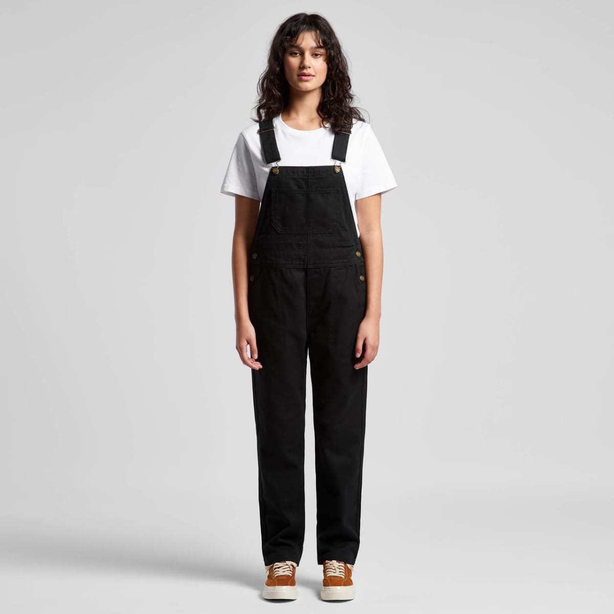 Person in black overalls, white t-shirt, and brown shoes standing against a plain background.