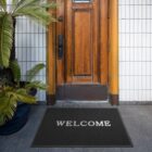 A doormat with WELCOME in front of a wooden door, next to a potted palm plant.