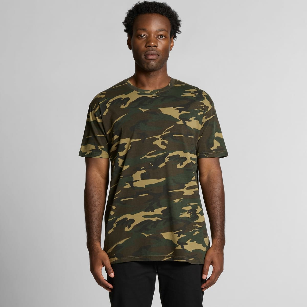Man wearing a green camouflage t-shirt and black pants stands facing forward against a gray background.