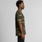 Man standing sideways wearing a green camouflage t-shirt and black pants on a plain background.